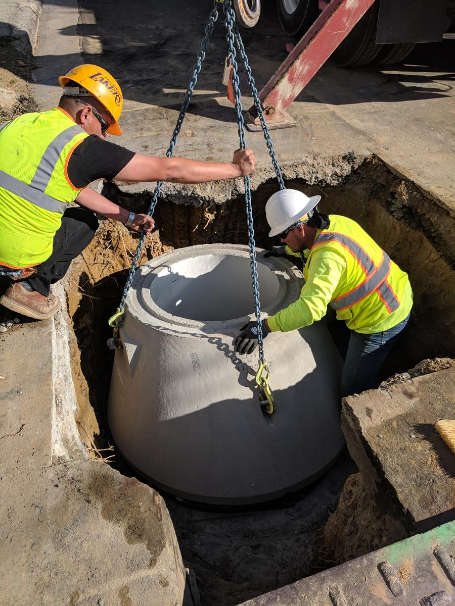 Two workers in hard hats and high-visibility vests rigging a precast concrete manhole section into an excavation