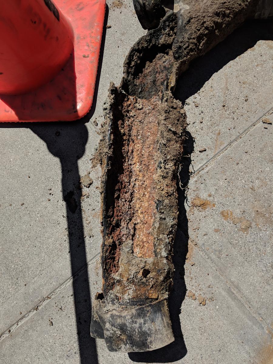 Section of badly corroded cast iron sewer pipe pulled from the ground, next to an orange traffic cone