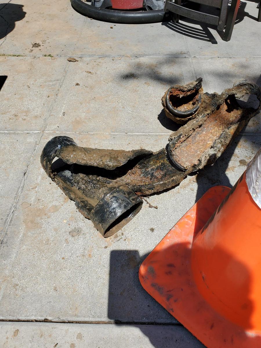 Old cast iron sewer fitting heavy with rust and scale, lying on pavement next to an orange cone