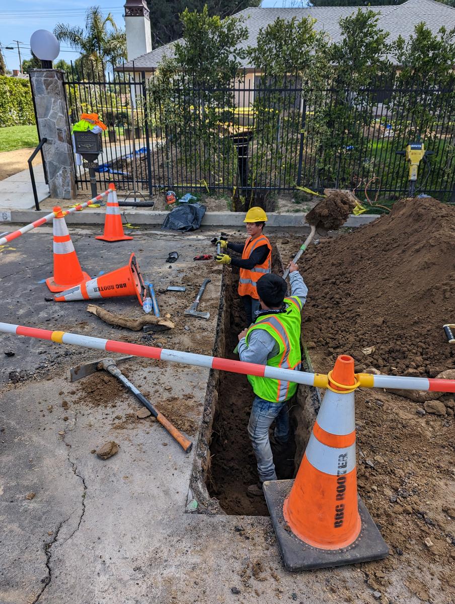 Crew in high-visibility vests working an open sewer trench behind traffic cones and caution tape