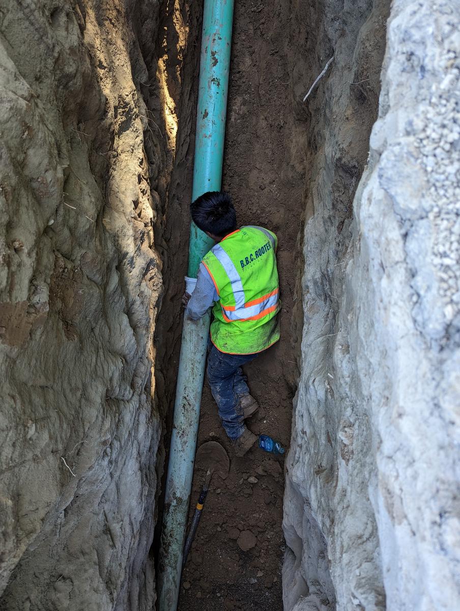 Crew member in a high-visibility vest working in a deep shored trench beside a sewer pipe