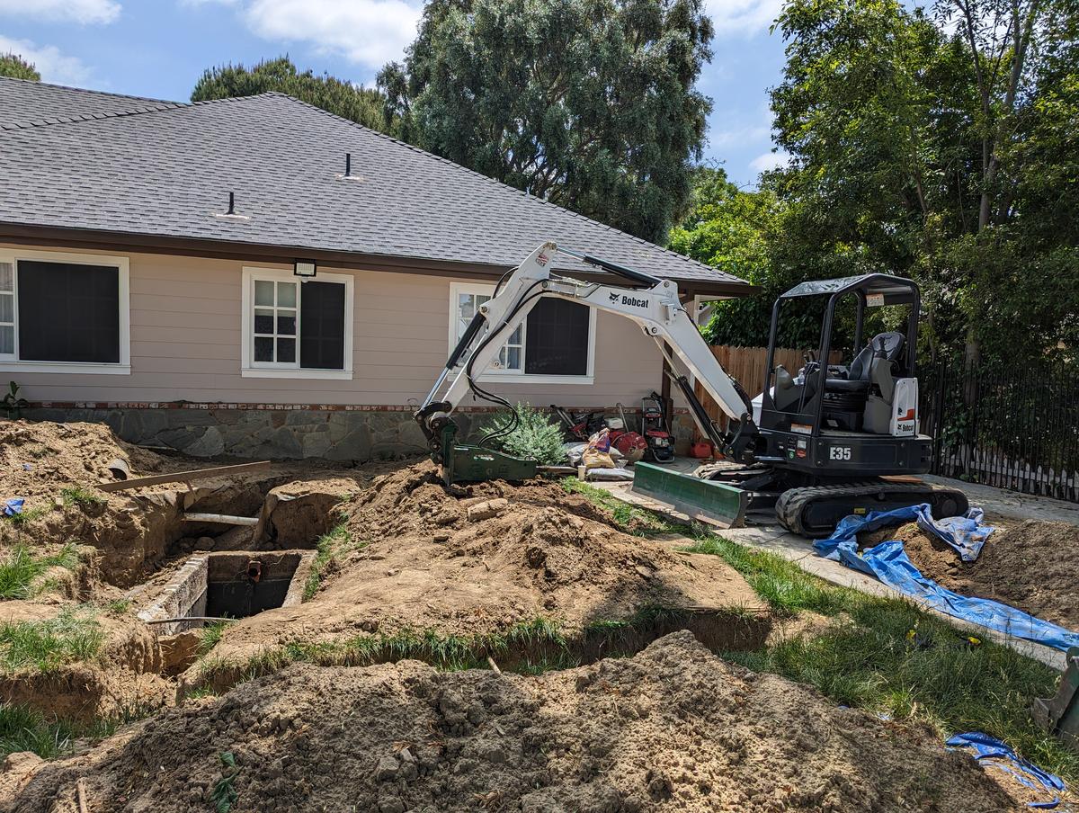 Compact excavator beside an open sewer trench and access pit next to a stucco building
