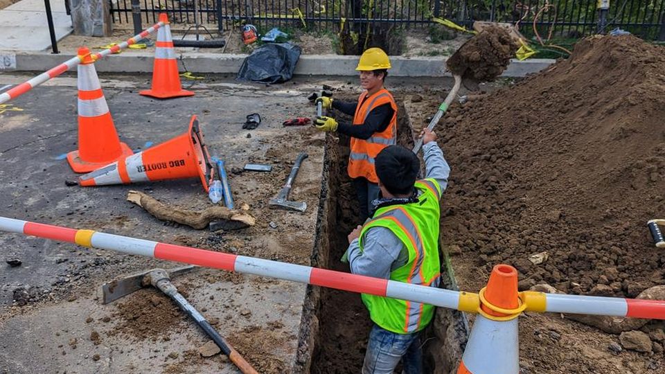 BBC Rooter crew working an open sewer trench behind traffic cones and caution tape